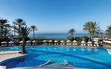 Großer Pool mit Palmen und Sonnenliegen, dahinter eine weitläufige Rasenfläche und der Blick auf das ruhige Meer.