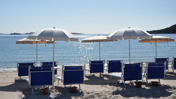 Reihe stilvoller blauer Liegestühle mit weißen Sonnenschirmen am ruhigen Sandstrand – mit Blick auf das glitzernde Meer und ferne Inseln unter klarem Himmel.