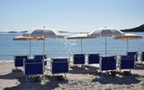 Reihe stilvoller blauer Liegestühle mit weißen Sonnenschirmen am ruhigen Sandstrand – mit Blick auf das glitzernde Meer und ferne Inseln unter klarem Himmel.