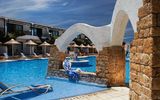 Einladender Poolbereich mit stilvollen Steinbögen, Sonnenliegen und strohgedeckten Sonnenschirmen vor zweistöckigen Hotelgebäuden – perfekte Kulisse für entspannte Urlaubstage unter strahlend blauem Himmel.