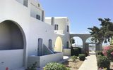 Ein gepflegter Gartenweg führt durch die hellen Gebäude im typisch kykladischen Stil mit weißen Fassaden und grauen Fensterläden – im Hintergrund lädt der Poolbereich zum Entspannen ein.