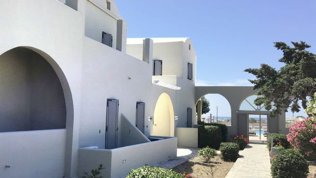 Ein gepflegter Gartenweg führt durch die hellen Gebäude im typisch kykladischen Stil mit weißen Fassaden und grauen Fensterläden – im Hintergrund lädt der Poolbereich zum Entspannen ein.