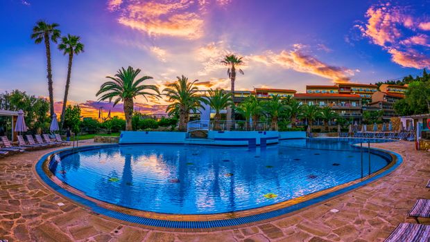 Großer Hotelpool mit Palmenkulisse bei Sonnenuntergang – das Wasser spiegelt den farbenprächtigen Himmel, umrahmt von Liegestühlen und mediterranem Ambiente.