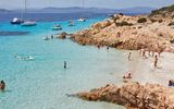 Traumhafter Strand mit türkisfarbenem Wasser, weißen Sandbuchten und badenden Urlaubern – umgeben von Felsen und Booten vor malerischer Küstenkulisse.