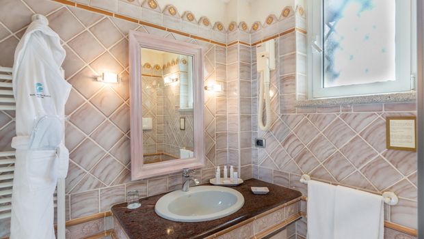 Stilvoll gefliestes Badezimmer mit Waschtisch, großem Spiegel, Kosmetikartikeln, Handtüchern, Bademantel und Tageslichtfenster – hell und gepflegt.