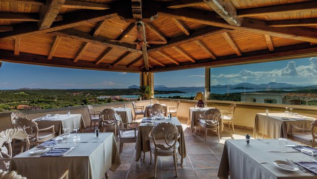 Elegantes Restaurant mit Holzdach und Panoramafenstern – gedeckte Tische mit Blick auf das Meer, grüne Landschaften und ferne Inseln unter blauem Himmel.