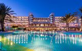 Beleuchtetes Hotel mit großem Poolbereich bei Nacht, eingerahmt von Palmen und klarer Abendstimmung.