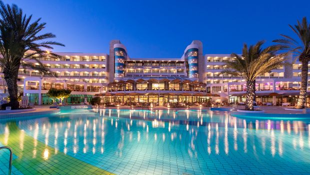 Beleuchtetes Hotel mit großem Poolbereich bei Nacht, eingerahmt von Palmen und klarer Abendstimmung.